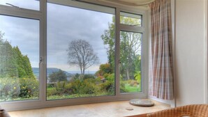 A room with a large window offering a view of a garden and hills. - West Ridge Bungalow (Seaton)