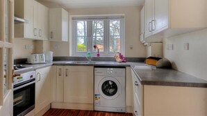 A compact kitchen with white cabinets, a built-in washing machine, a toaster, and a window with a view of greenery. - 4 Riverside Cottages (Bridport)
