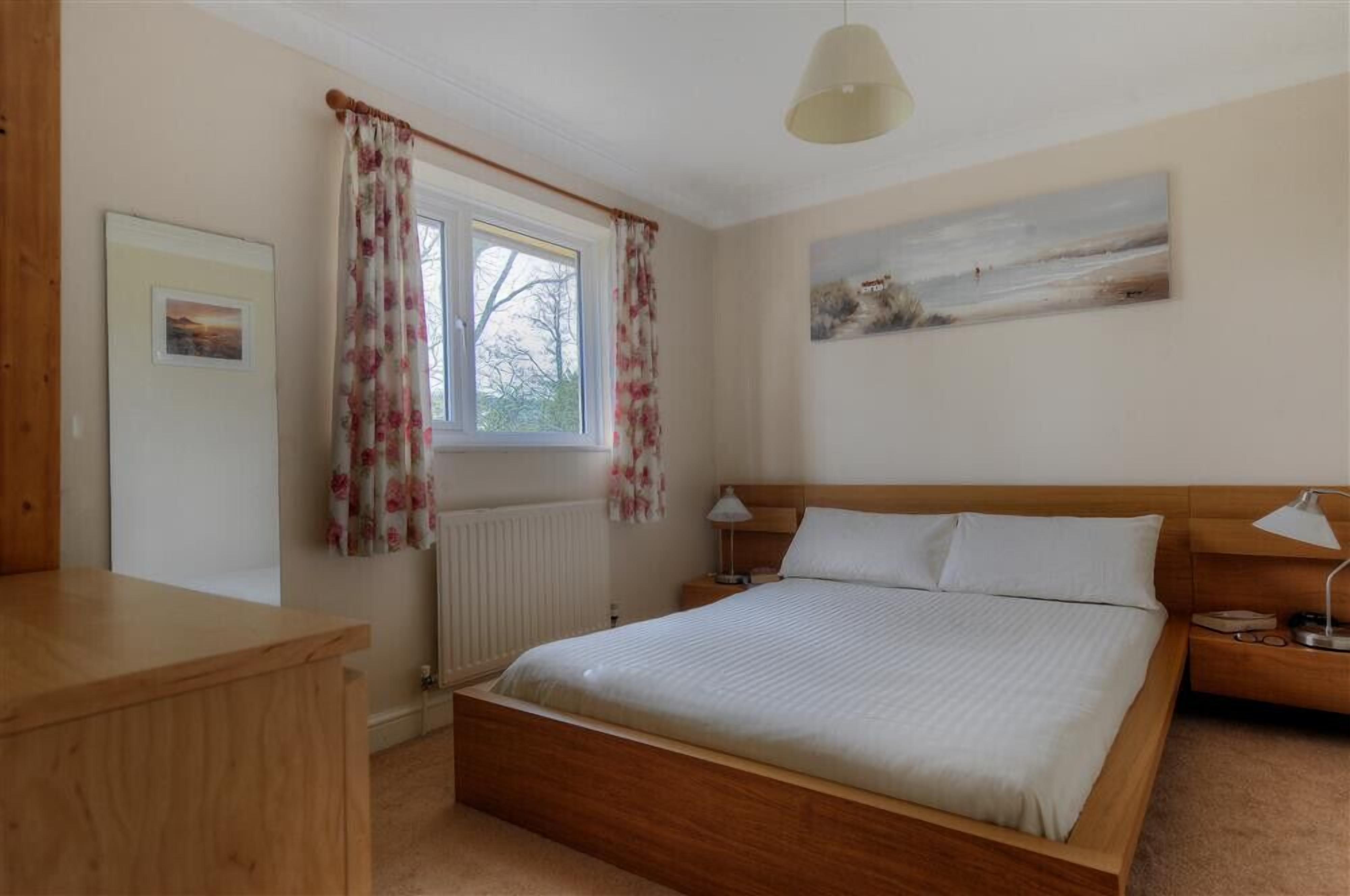 A bedroom with a wooden bed, a nightstand, a desk, a window with floral curtains, and a painting on the wall.