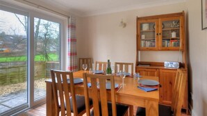 A dining area with a wooden table set for four, a cabinet with glass doors, and a view of the outdoors through a large window. - 4 Riverside Cottages (Bridport)