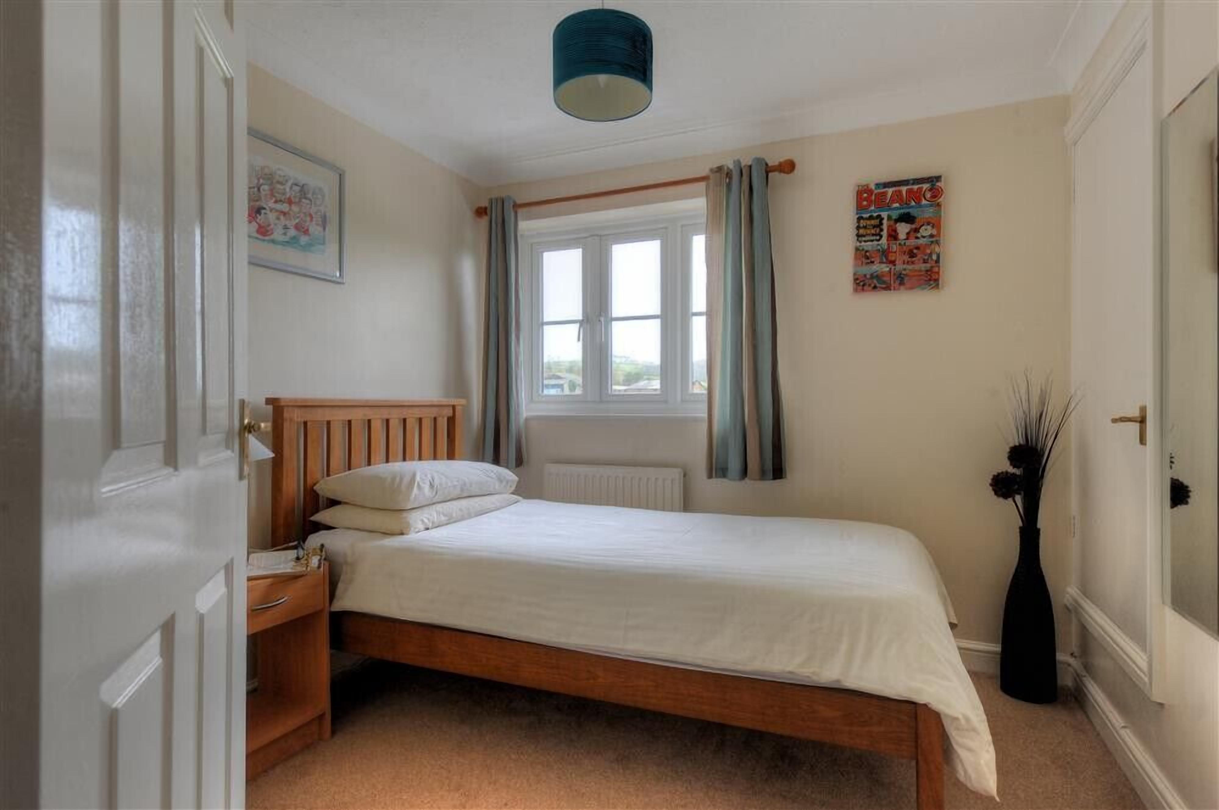 A bedroom with a wooden bed, a nightstand, a window with curtains, a radiator, and a framed picture on the wall.