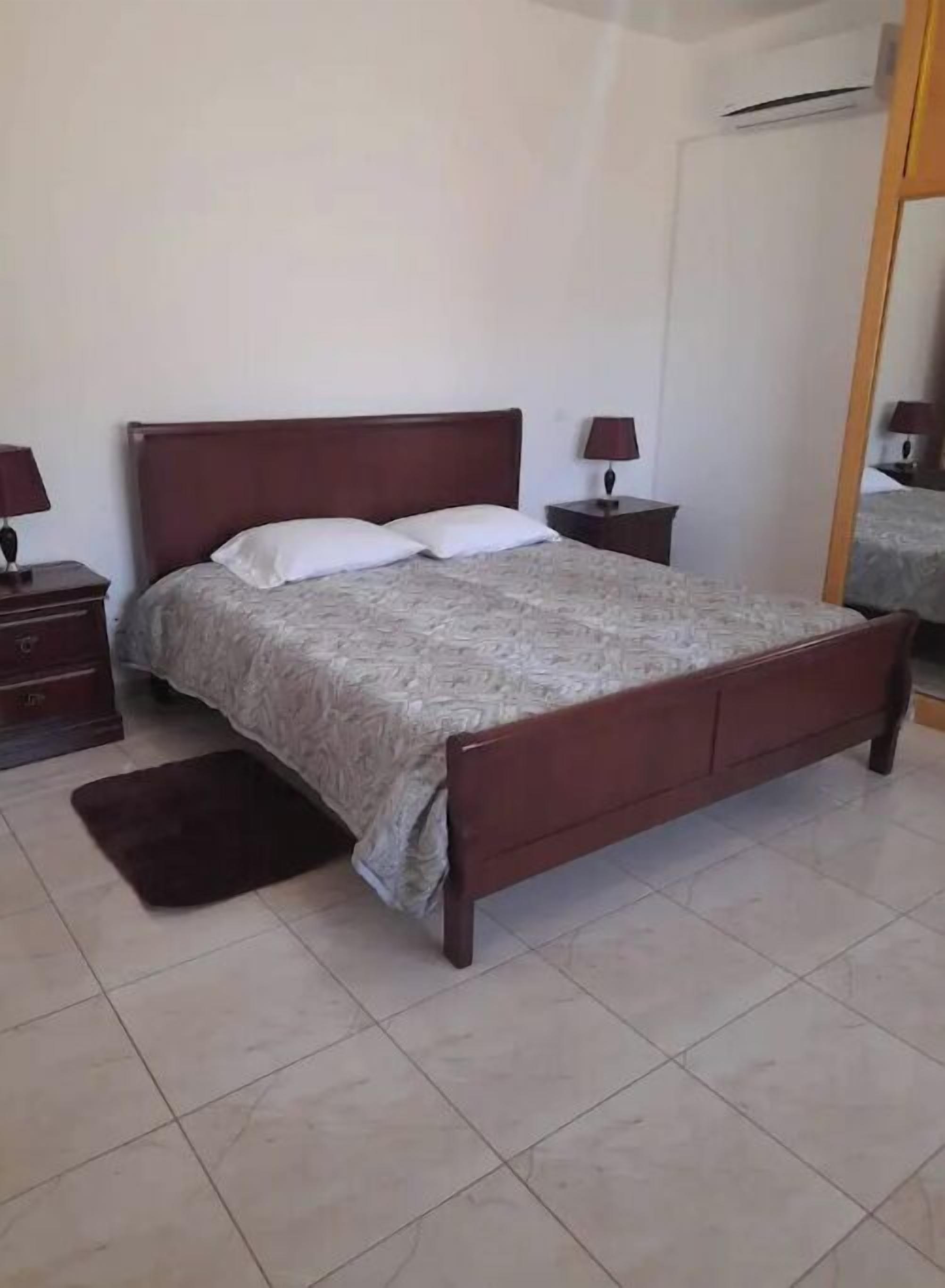 Un dormitorio con cama de madera, dos mesitas de noche con lámparas, alfombra oscura y suelo de baldosas.