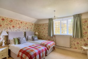 Une chambre avec un papier peint à motifs floraux, deux lits recouverts de couvre-lits à carreaux, un radiateur, une fenêtre avec des rideaux et une table de chevet avec une lampe.