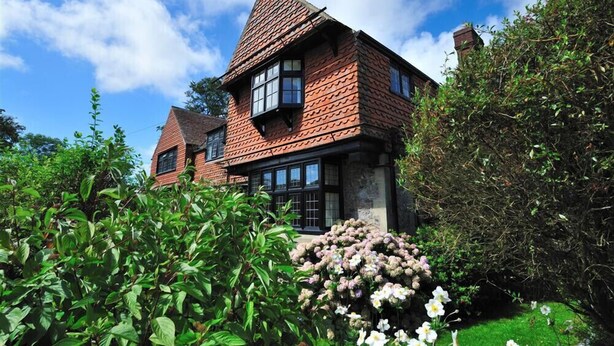 Une maison traditionnelle avec un toit à forte pente, de nombreuses fenêtres et un jardin bien entretenu, agrémenté de diverses plantes et fleurs.