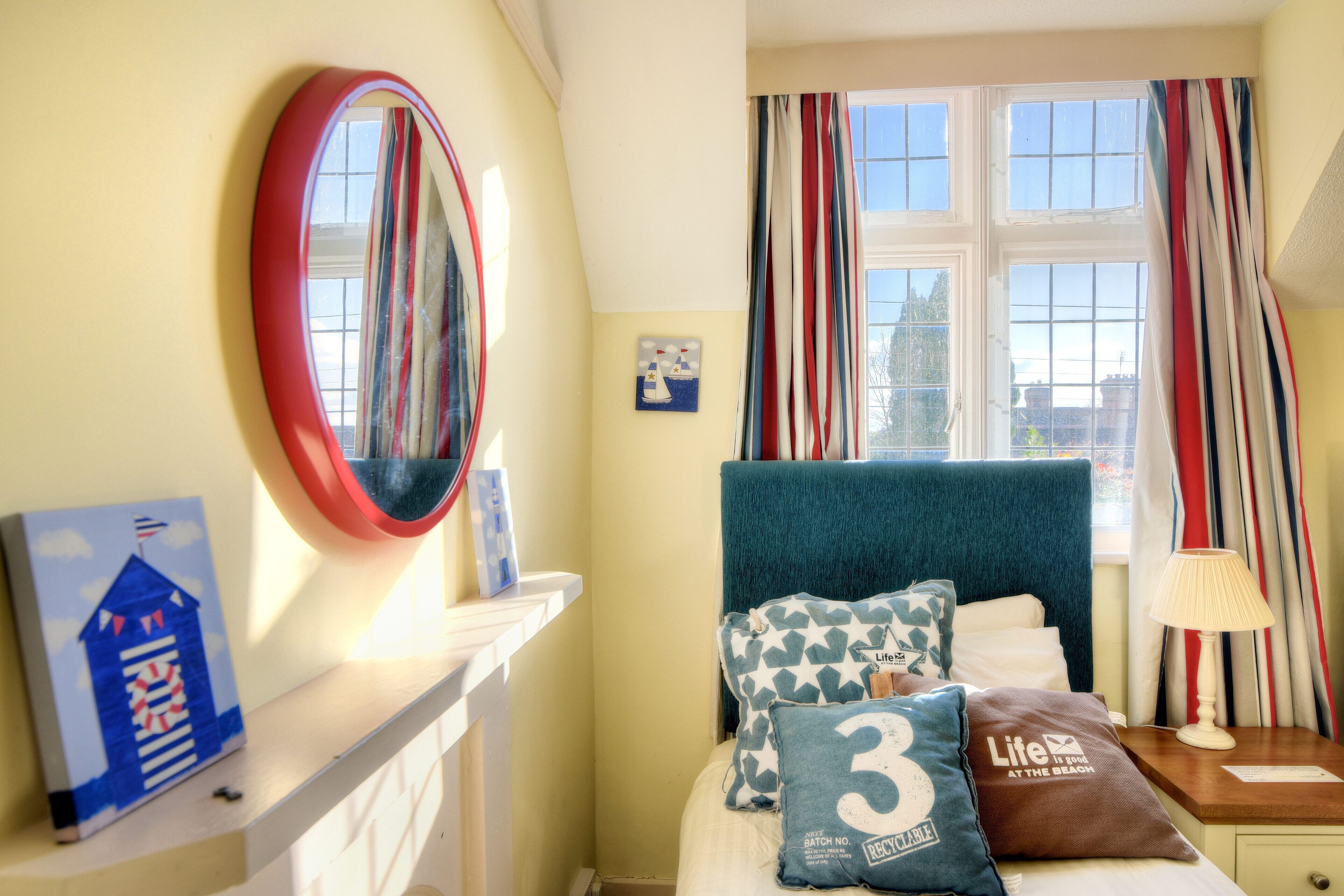 A bedroom with a bed, a bedside table with a lamp, a window with curtains, a mirror, and a small picture on the wall.