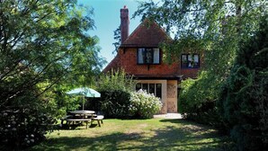 Une maison traditionnelle avec une cheminée en briques, entourée d’une végétation luxuriante et dotée d’un petit patio avec une table et des chaises.
