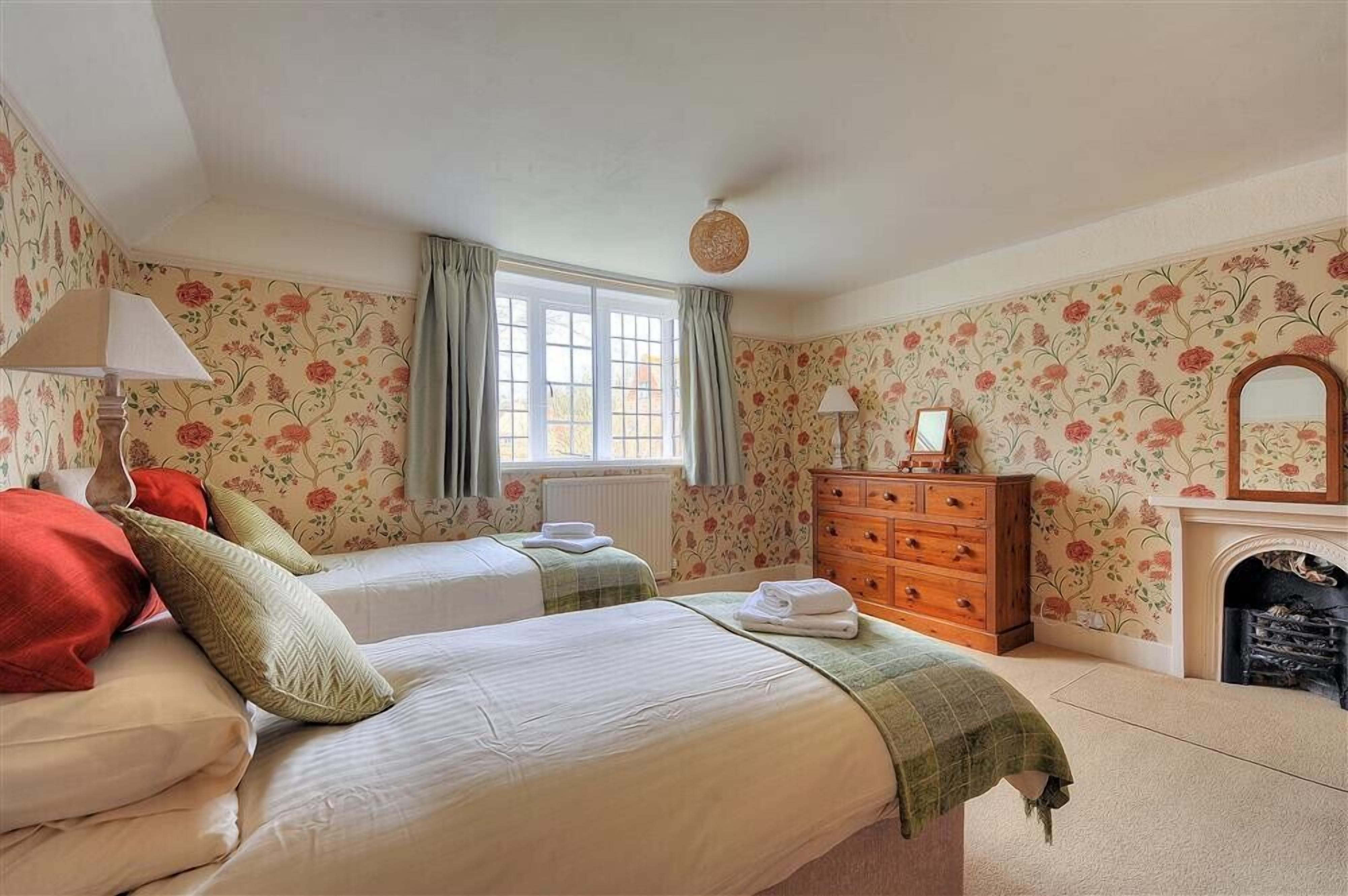 A bedroom with two beds, a dresser, a mirror, a fireplace, and floral wallpaper.