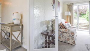 A room with a wooden desk, a lamp, a sofa with patterned cushions, and a large window with a view of greenery. - Byeways (Axminster)