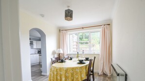 A dining area with a table set for four, a window with curtains, and a kitchen in the background. - Byeways (Axminster)
