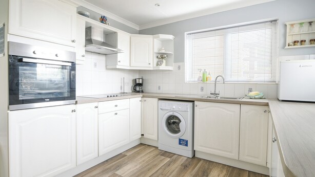 A modern kitchen with white cabinetry, a built-in oven, a washing machine, and a window with blinds. - Byeways (Axminster)