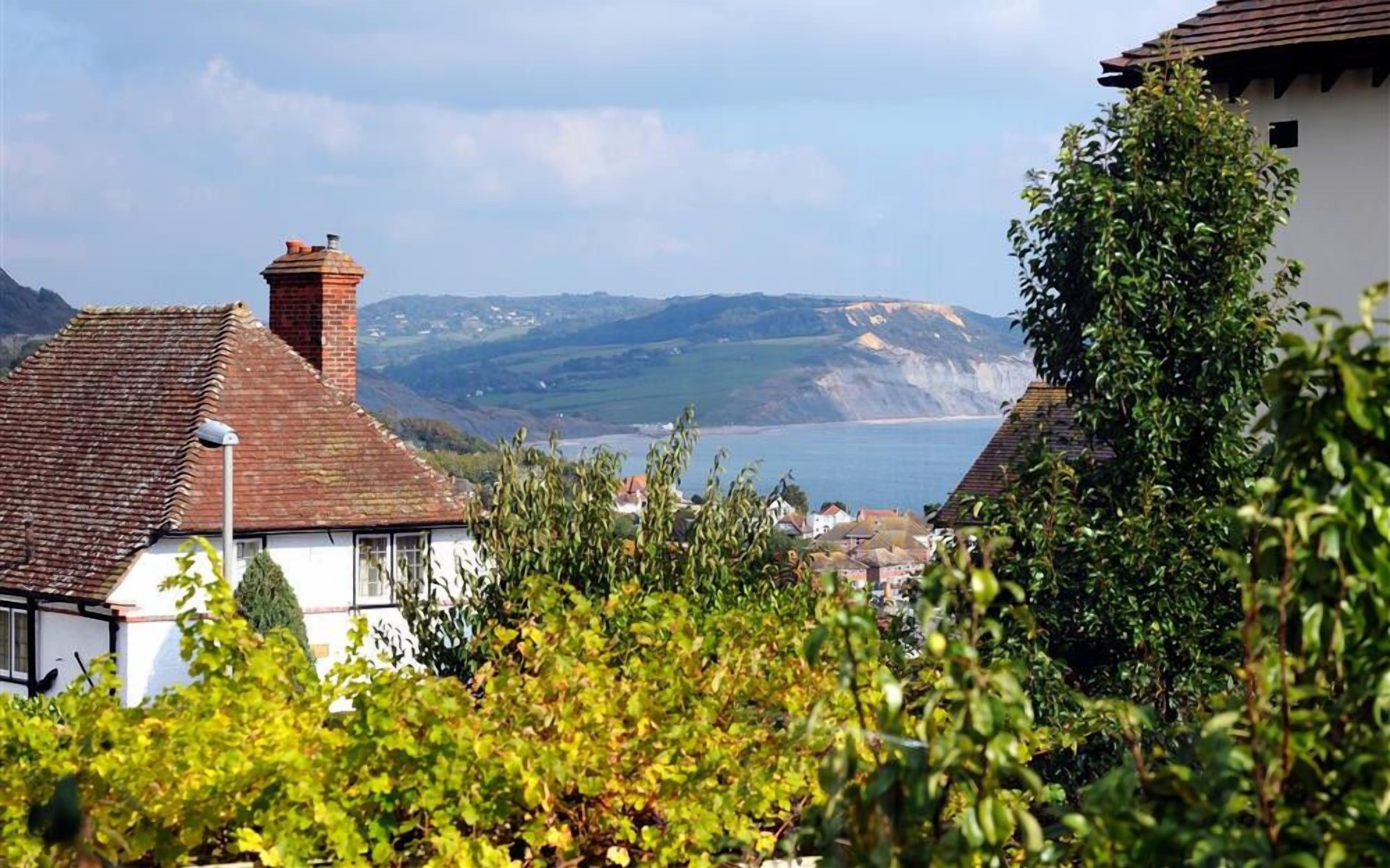 A coastal village with white houses, a view of the sea, and hills in the background.