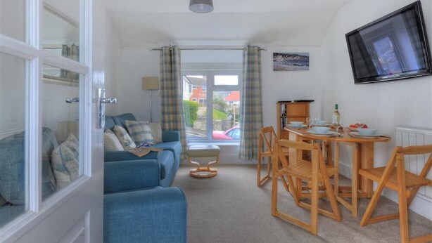 A living room with a blue sofa, wooden dining table, and chairs. There is a window with curtains, a television on the wall, and a small table with a lamp. - Hatchett Top Flat (Lyme Regis)