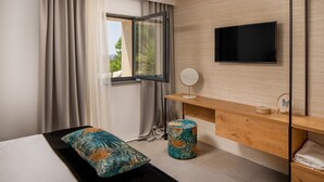 A hotel room with a bed, a wooden desk, a TV, a window with curtains, and a view of greenery outside. - Villa Maremonte with Pool (Kaštel Kambelovac)