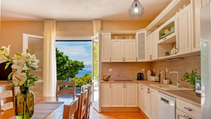 A modern kitchen with white cabinets, a dining area with a table and chairs, and a view of the sea through an open door. - Luxury Villa Aerius with Pool (Pučišća)
