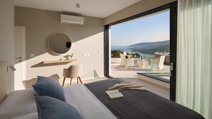 A bedroom with a large bed, a round mirror, a small table with a chair, and a view of a balcony with a hot tub and a scenic landscape.