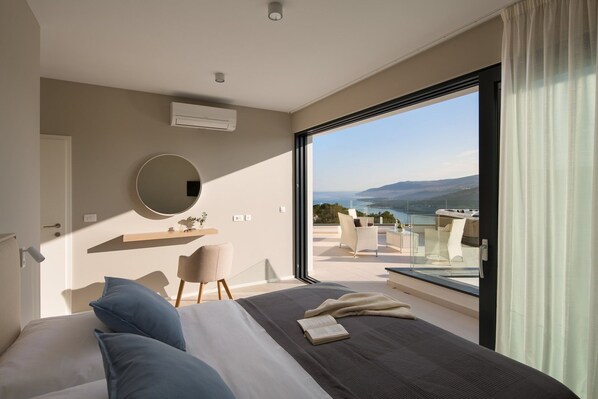 A bedroom with a large bed, a round mirror, a small table with a chair, and a view of a balcony with a hot tub and a scenic landscape.