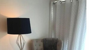 Una habitación con un sillón color beige, una mesita redonda blanca, una lámpara de pie y una cortina gris.