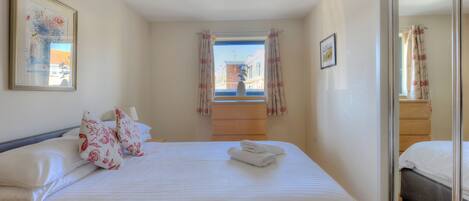 A neatly made bed with white linens and floral pillows, a window with curtains, and a framed picture on the wall.