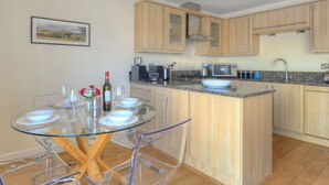 A modern kitchen with wooden cabinets, a granite countertop, and a glass dining table with chairs. - Harbour View (Bridport)