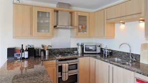 A modern kitchen with wooden cabinets, a granite countertop, a built-in oven, a microwave, and a sink. - Harbour View (Bridport)