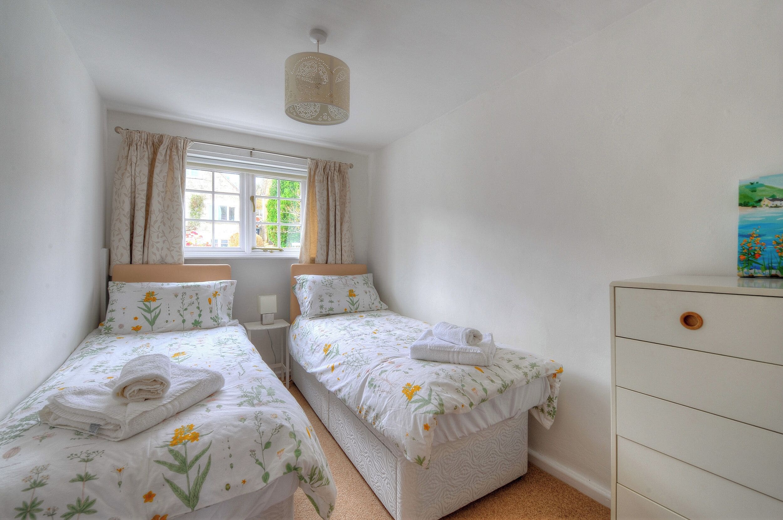 A bedroom with two single beds, a dresser, a window with curtains, and a painting on the wall.