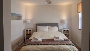 A neatly made bed with white linens and towels, two bedside tables with lamps, a painting on the wall, and a window with striped curtains. - Little Ascot (Lyme Regis)