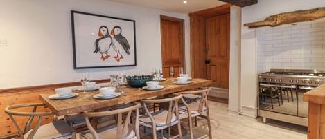 Zona pranzo con tavolo di legno apparecchiato per quattro, circondato da sedie in vimini. Sopra il tavolo, un quadro raffigura uccelli.