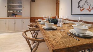 A dining area with a wooden table set for four, surrounded by chairs. There is a kitchen area with white cabinets and a shelf with dishes. A framed artwork of puffins is on the wall. - Captains House (Lyme Regis)