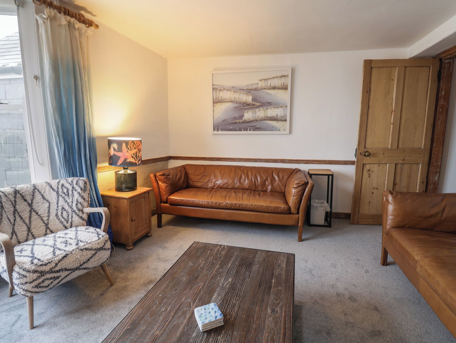 Una sala de estar con un sofá de cuero marrón, un sillón estampado y una mesa de centro de madera. Un cuadro cuelga en la pared y se ve una puerta de madera.