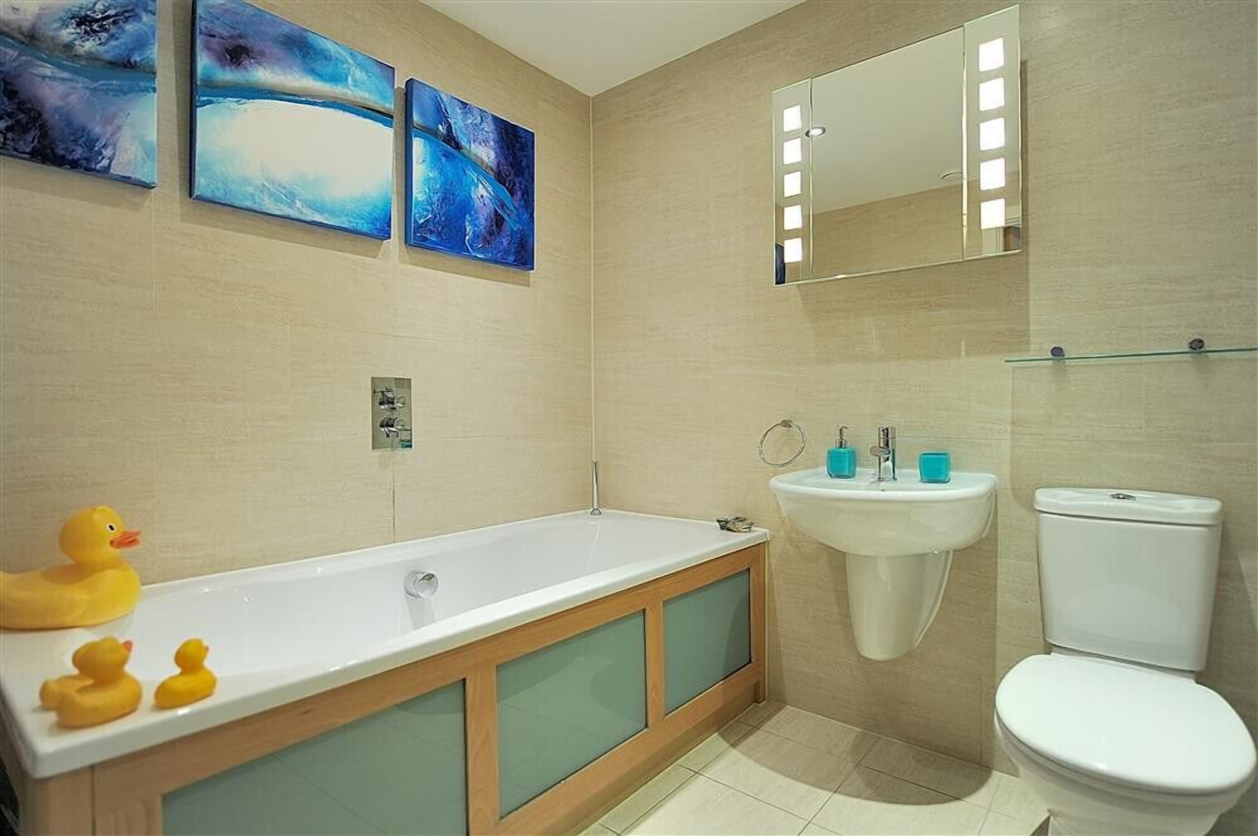 Un baño con bañera, lavamanos e inodoro. Hay tres cuadros en la pared y un patito de goma en la bañera.