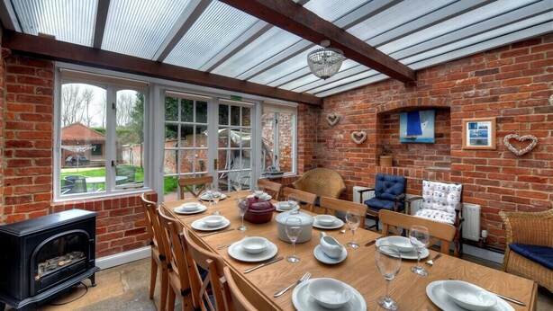 A dining area with a brick wall, a wooden table set for four, and a black wood stove. - The White House (Bridport)