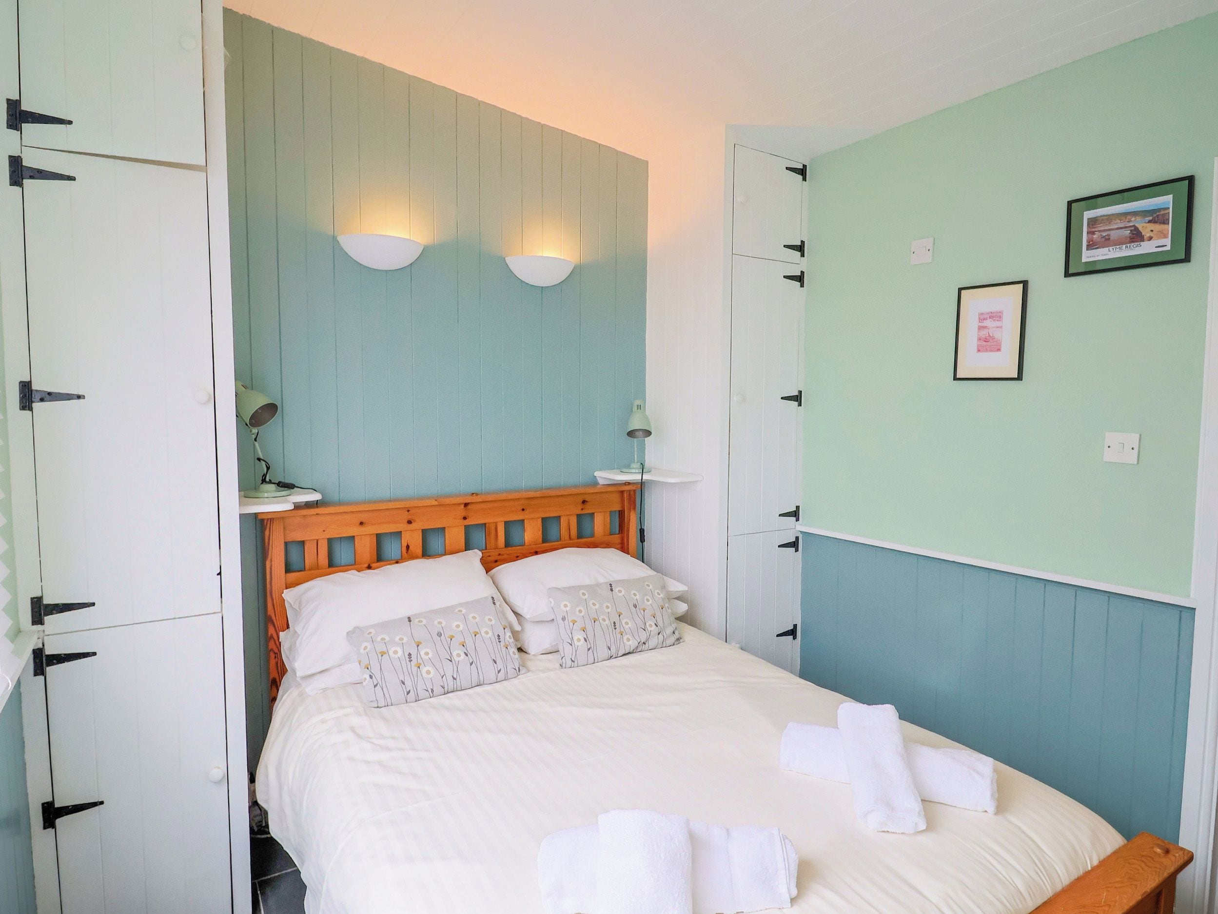 Un dormitorio con cama, almohadas, mesita de noche, lámparas de pared y un cuadro en la pared.