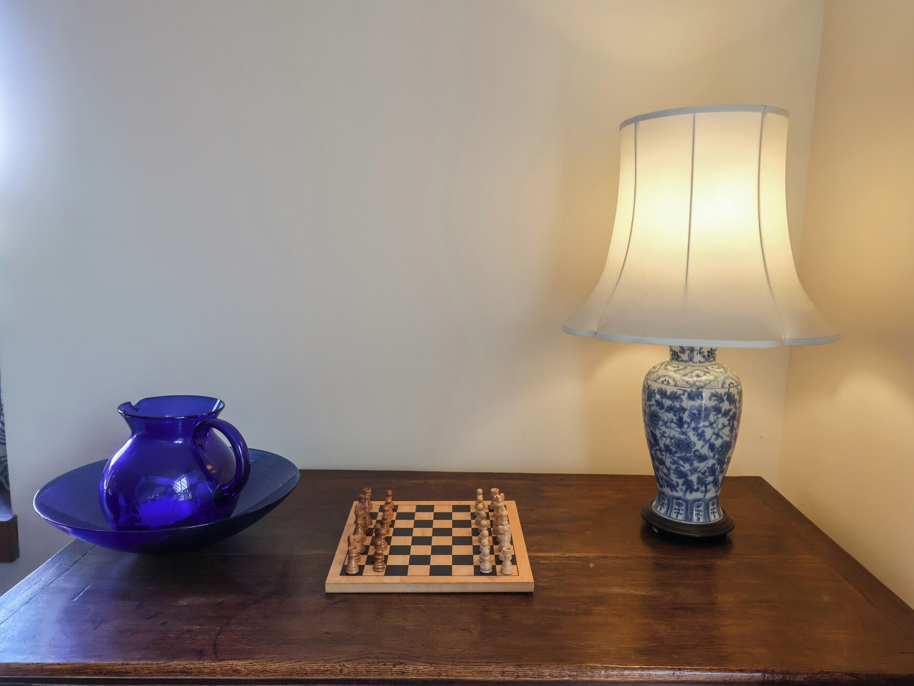 Une table en bois avec un jeu d’échecs, un vase bleu et une lampe.