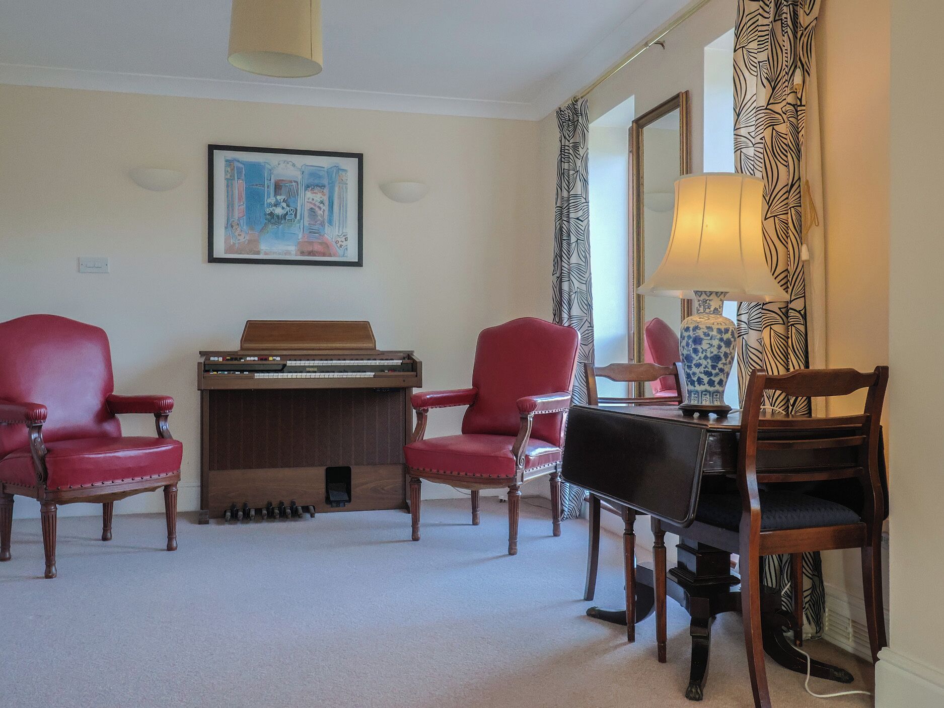 A room with a piano, two red chairs, a table with a lamp, a framed picture on the wall, and patterned curtains.