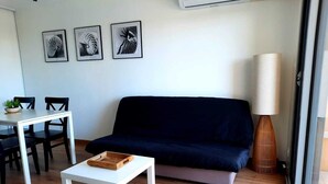 Een woonkamer met een donkerblauwe bank, een witte salontafel, een eettafel met stoelen en drie ingelijste foto's aan de muur.
