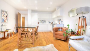 A modern kitchen with white cabinets, a dining area with wooden chairs, and a living room with a sofa and a lamp. - Lyme Time (Lyme Regis)