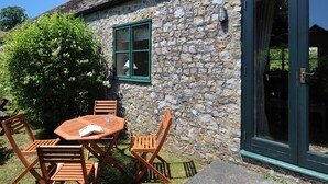 A stone building with a green door and window, outdoor seating with a table and chairs, and a bush with white flowers. - 3 Home Farm South (Lyme Regis)