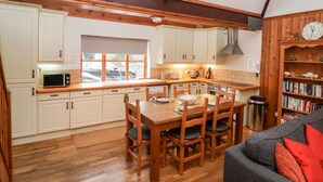 A kitchen with wooden cabinets, a dining table, chairs, a microwave, and a bookshelf. - 3 Home Farm South (Lyme Regis)