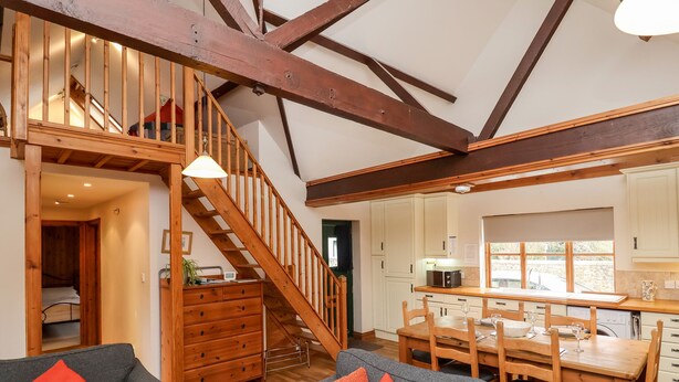 A spacious living area with a staircase, wooden beams, a dining table, and a kitchen area. - 3 Home Farm South (Lyme Regis)
