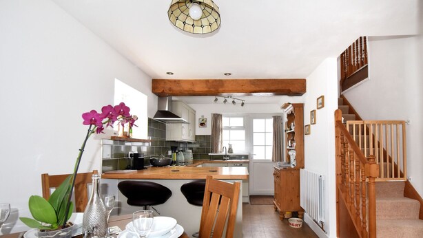 A modern kitchen with a wooden island, a dining area with a table set for two, and a staircase with wooden railings. - Sunnyside (Colyton)