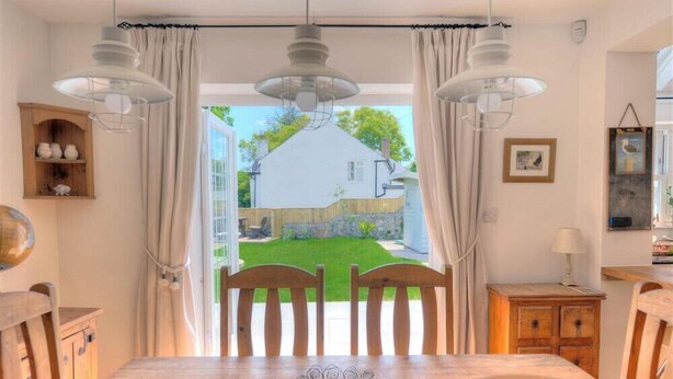 A dining area with a table and chairs, a wooden cabinet, and a view of a white house with a garden. - Bethel Cottage (Lyme Regis)