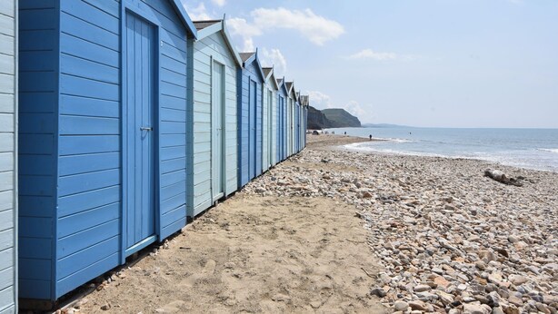 A row of blue beach huts along a pebbled beach with the sea in the background. - 44 Fernhill Heights (Bridport)