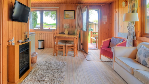 A cozy cabin interior with a wooden interior, a television mounted on the wall, a dining area with a table and chairs, and a sofa. - 4 Pinewood Retreat (Axminster)