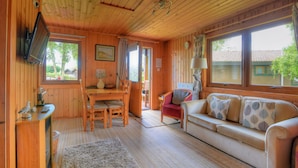 A cozy cabin interior with a wooden ceiling, a television mounted on the wall, a dining area with a table and chairs, a sofa with patterned cushions, and large windows offering a view of the outside. - 4 Pinewood Retreat (Axminster)