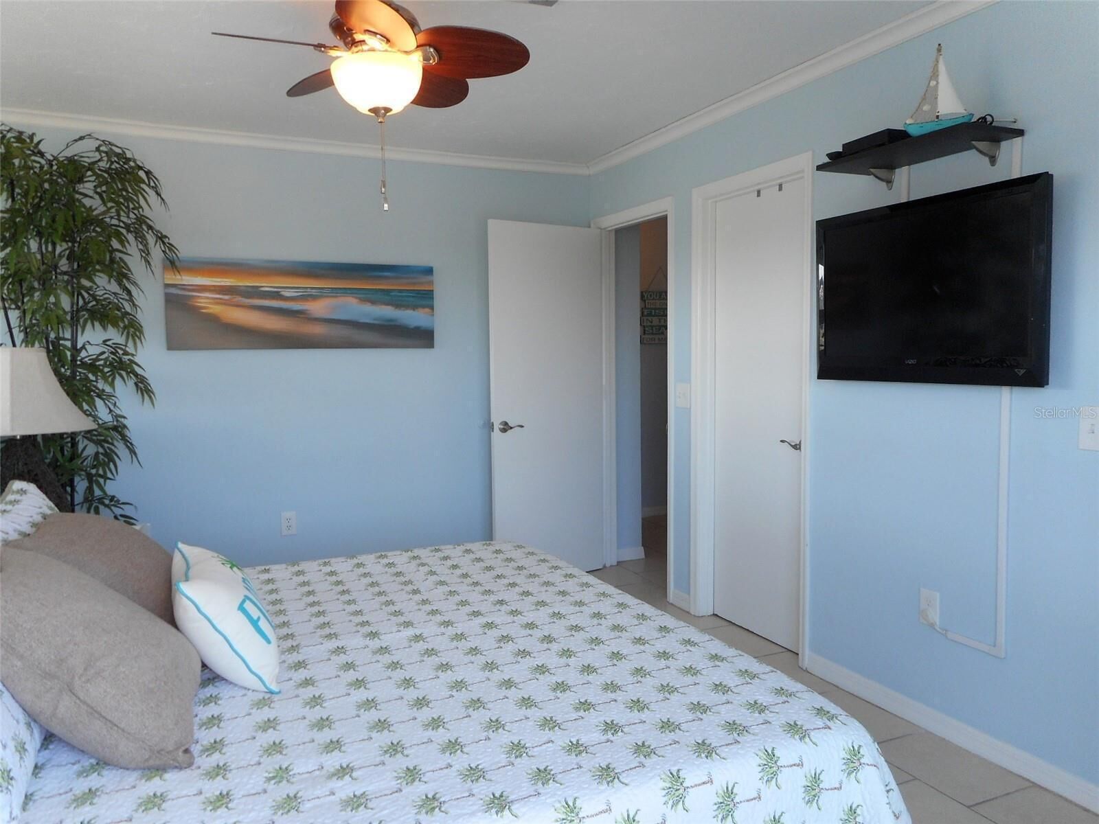 Een slaapkamer met een bed, een plafondventilator, een aan de muur bevestigde televisie en een deur naar een andere kamer.