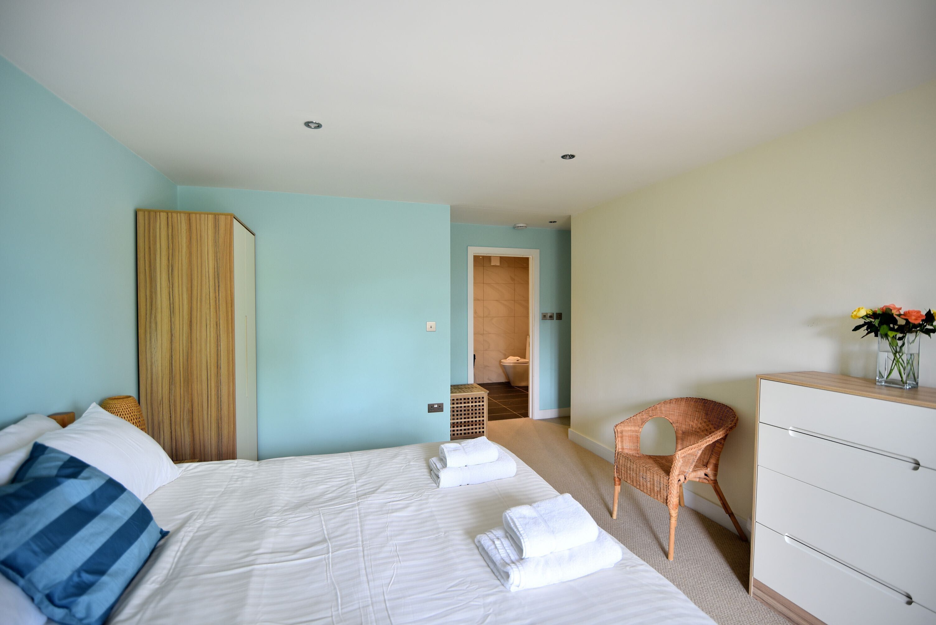 A hotel room with two beds, a wooden headboard, a wicker chair, a white dresser, and a bathroom visible through an open door.