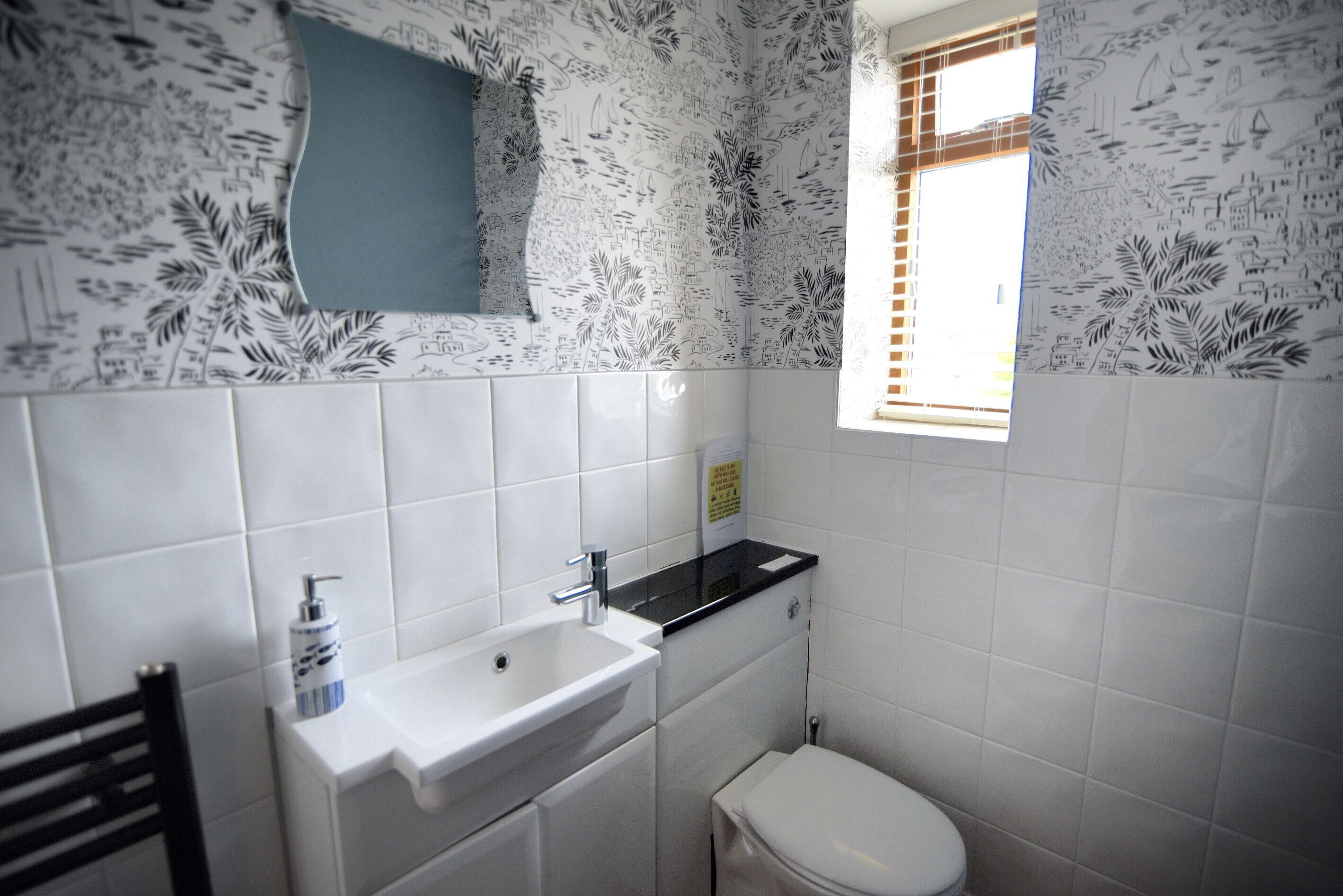 A bathroom with white tiles, a sink, a toilet, and a window with blinds.