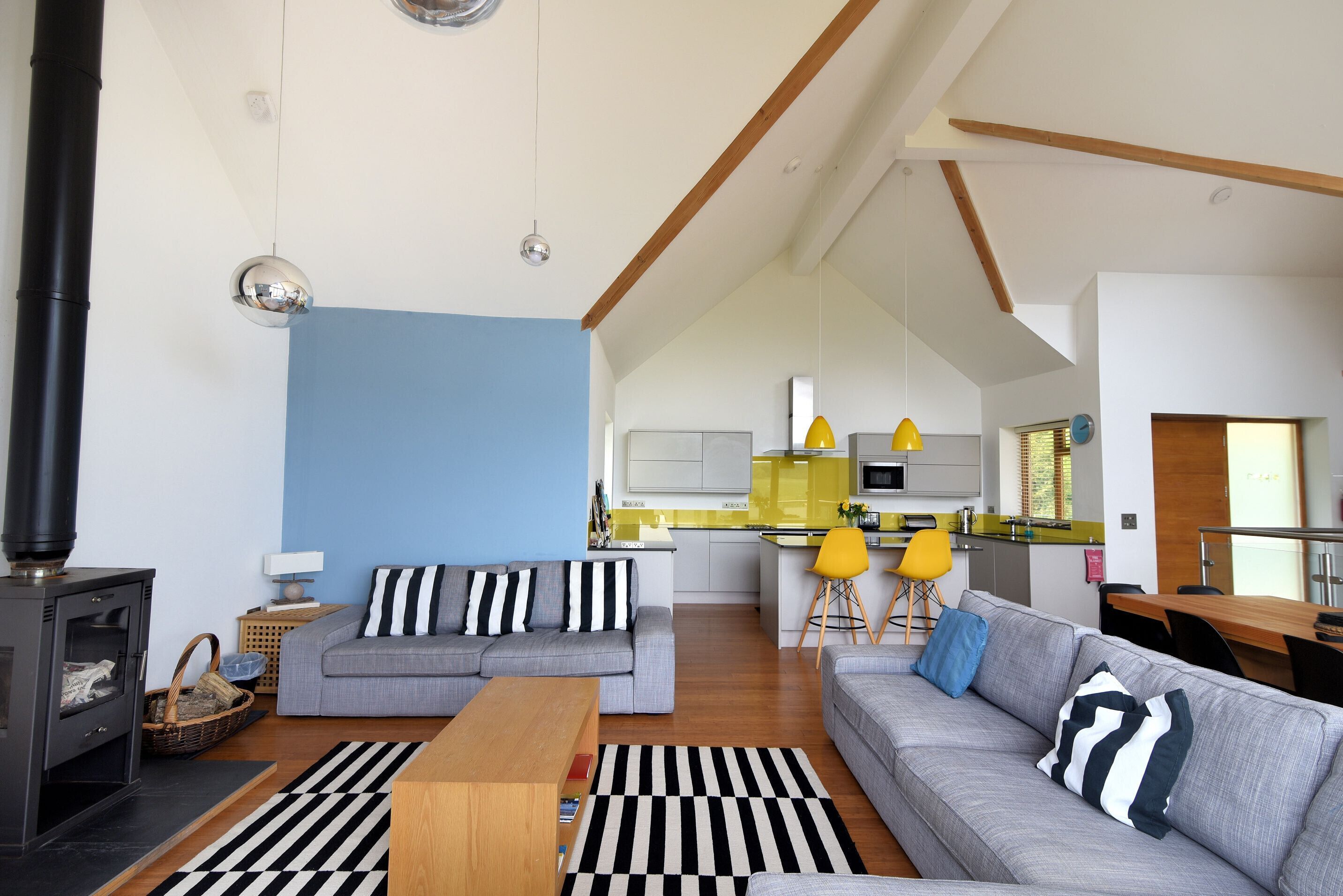 Ein modernes Wohnzimmer mit einer Couch, einem Holztisch und einem schwarz-weiß gestreiften Teppich. Im Hintergrund ist eine Küche mit gelben Stühlen und einer blauen Wand zu sehen.