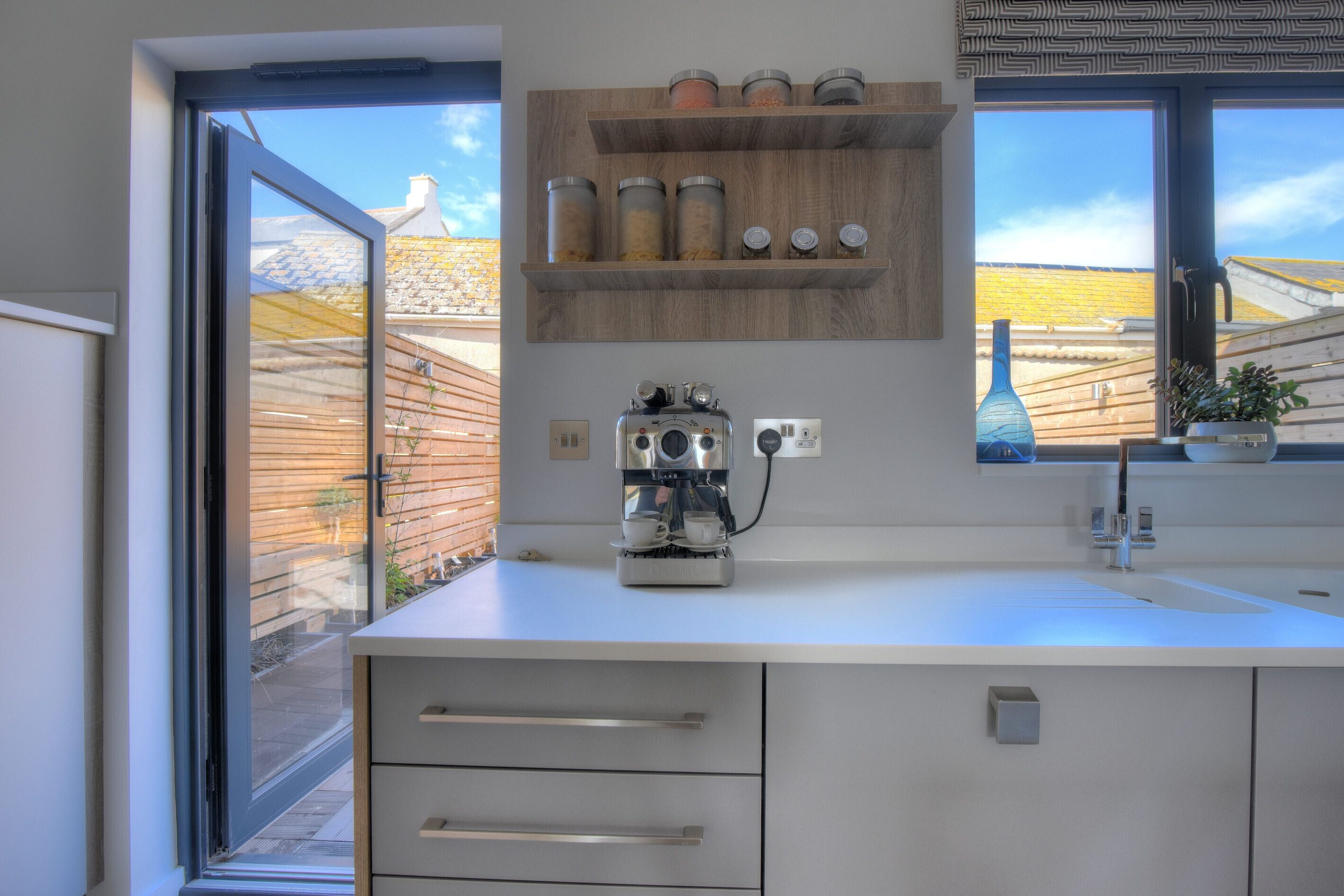 A modern kitchen with a built-in coffee machine, a window with a view of a yellow-roofed building, and a wooden shelf with jars.