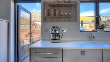 A modern kitchen with a built-in coffee machine, a window with a view of a yellow-roofed building, and a wooden shelf with jars.